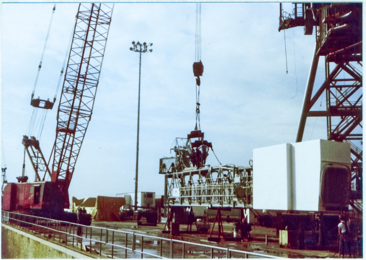 Union ironworkers attach a pair of spreader-beams to lifting lugs located on the top truss of the Orbiter Access Arm, which is still resting on its supports, on the pad deck at Space Shuttle Launch Complex 39-B, Kennedy Space Center, Florida, just prior to being lifted and attached to the Fixed Service Structure, where it will provide access for flight crew to enter the orbiter before it is launched into space.